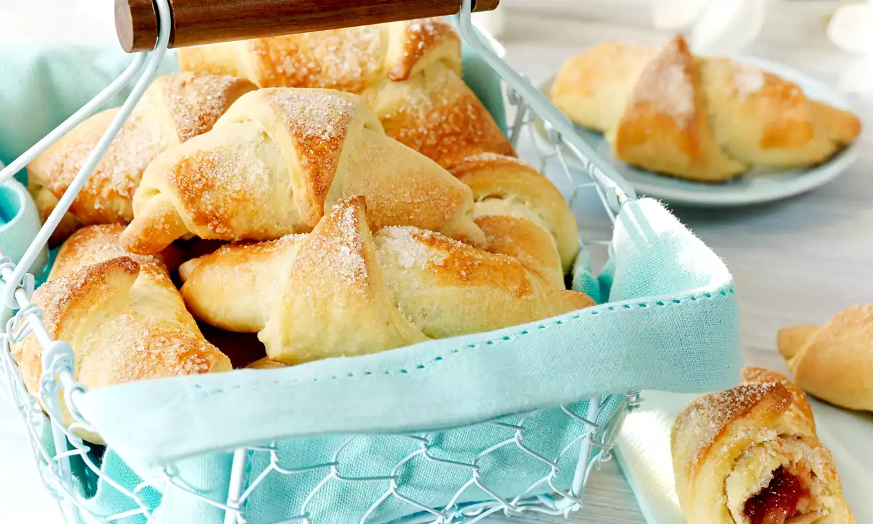 Złociste, półksiężycowe rogaliki drożdżowe z cukrową posypką ułożone w metalowym koszyku wyściełanym błękitnym materiałem na jasnym tle